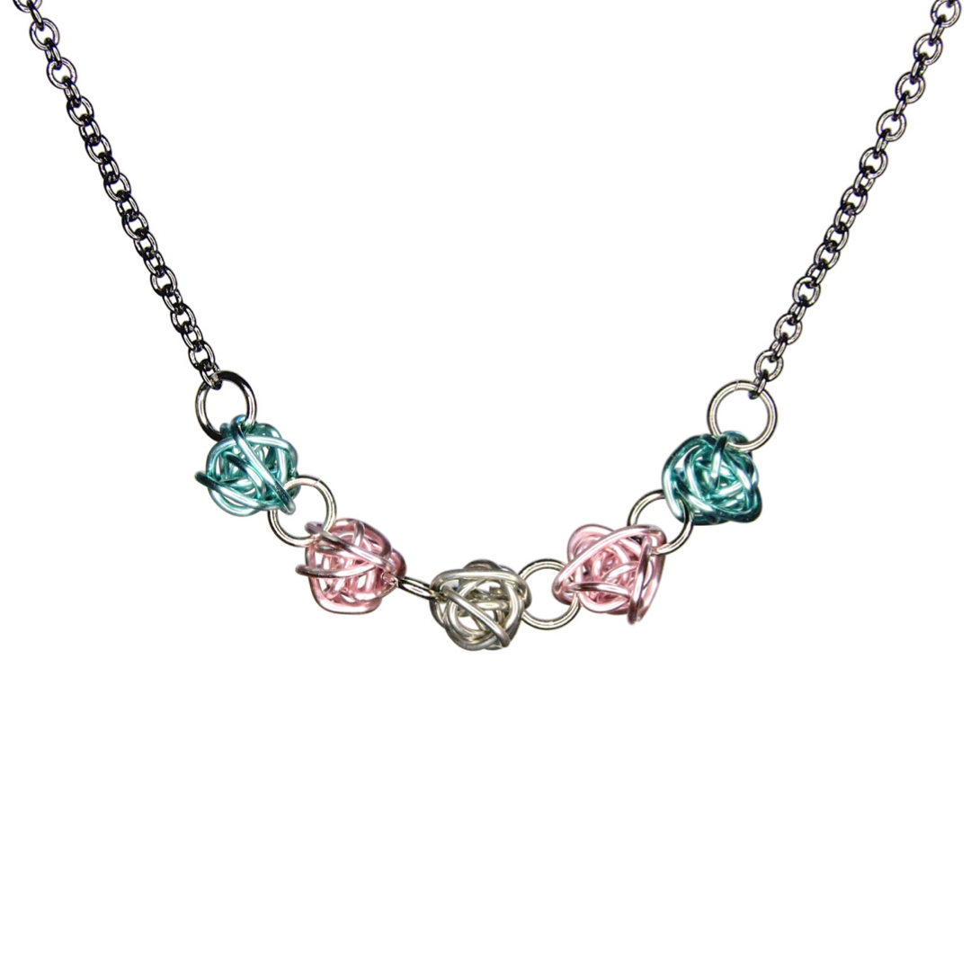 Necklace has 5 colorful loosely wrapped wire balls attached horizontally by jump rings in order of the trans pride flag - light blue, pink, silver, pink, light blue. The wire is shiny and smooth.