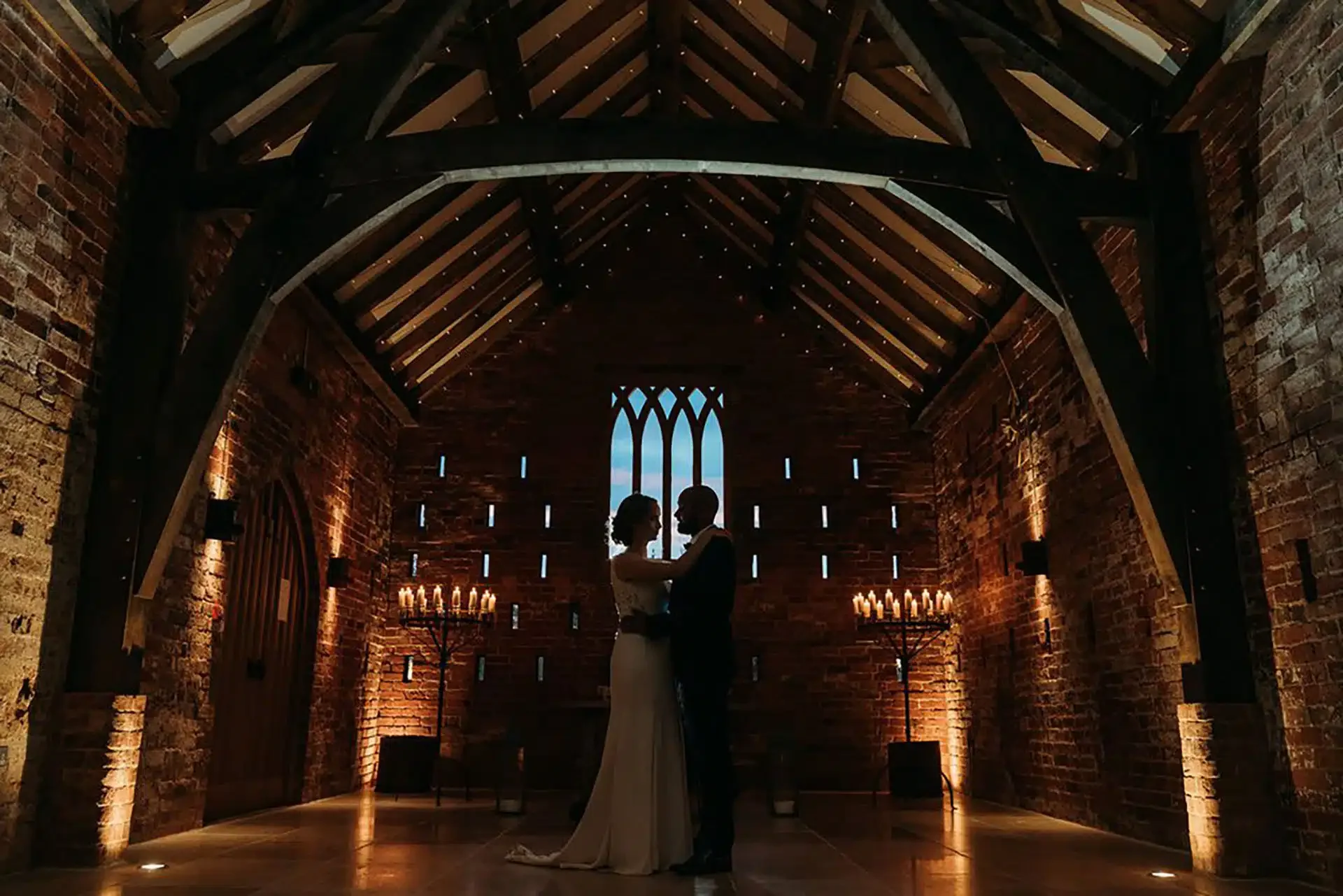 Silhouetted couple, bride and groom, standing close together inside Grangefields wedding venue with brick walls and wooden beams, candlelit candelabras on either side, in a romantic, intimate moment.