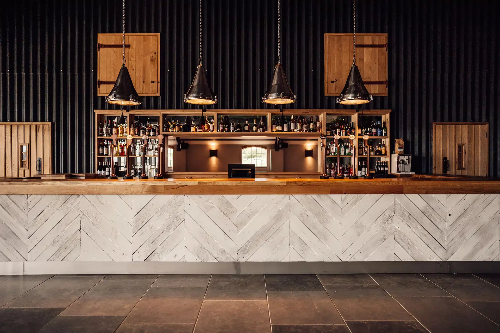 Grangefields wedding venue  bar counter with shelves of alcohol bottles, wooden panels, and black vertical wall decor. Hanging black pendant lights above the counter.