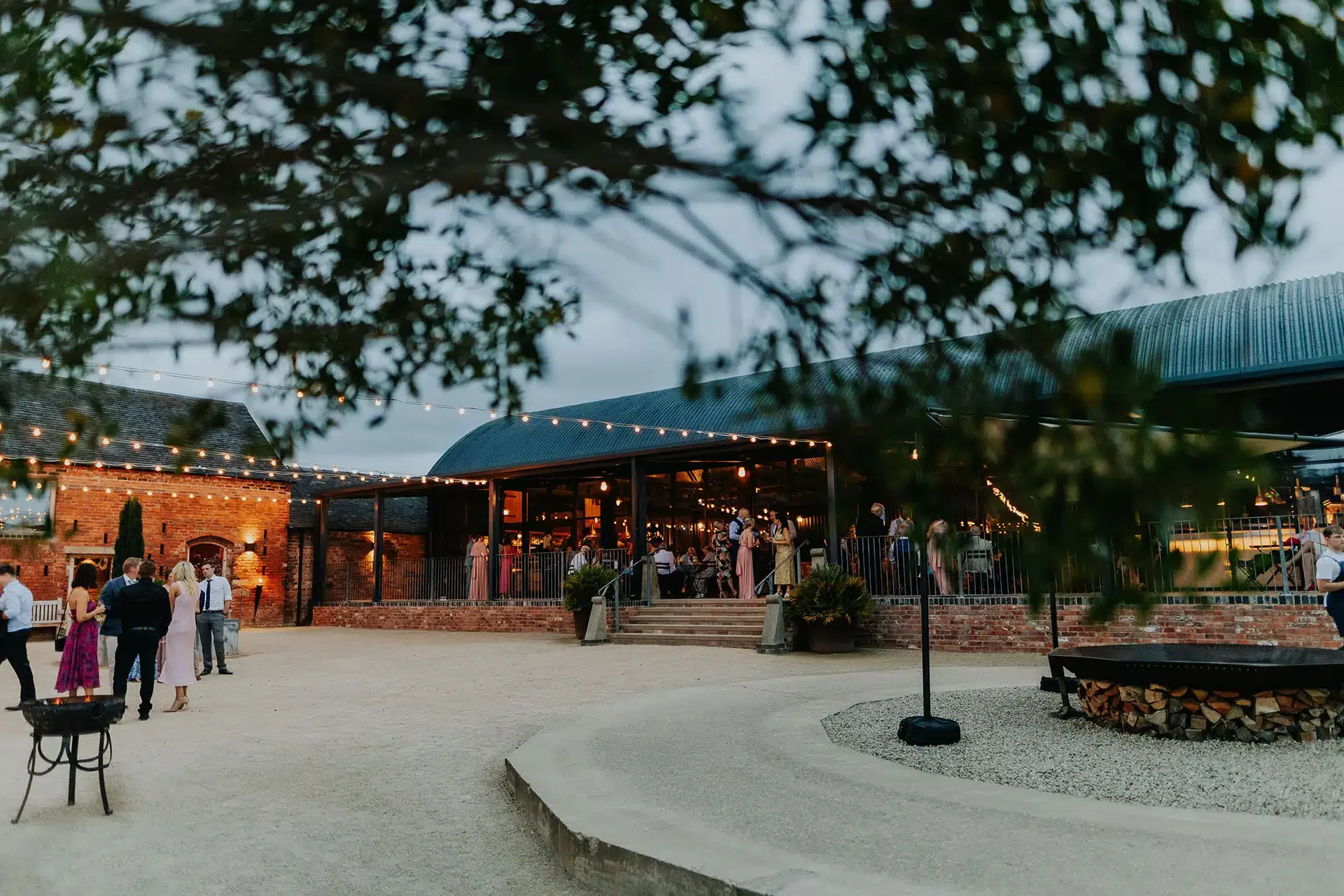 Outdoor social event at Grangefields wedding venue with string lights, people mingling on a gravel surface, brick building, and covered patio area.