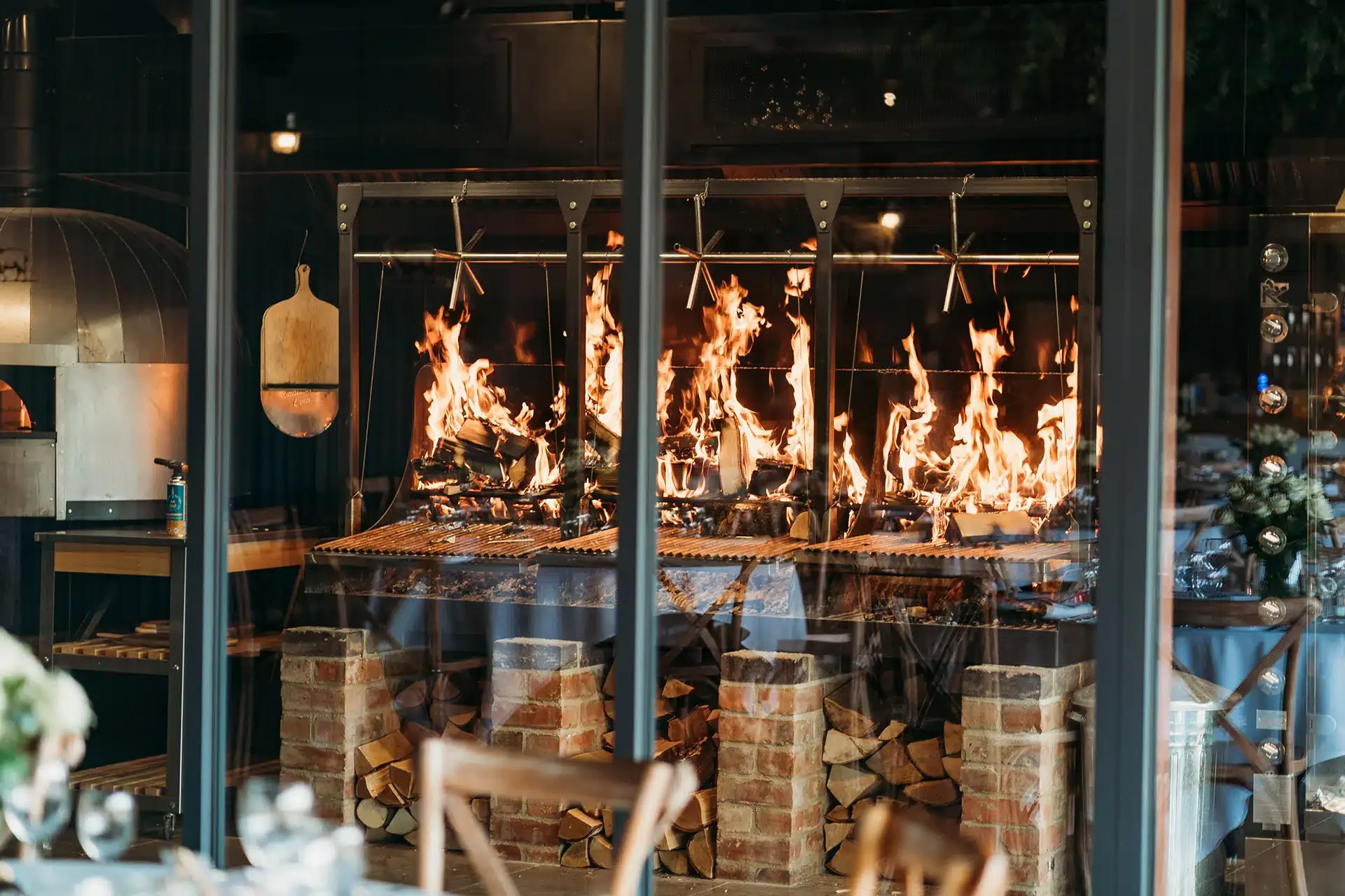 Grangefields wedding venue kitchen with a large open grill area on fire, with flames rising from stacked logs underneath a metal grill, enclosed by glass walls.