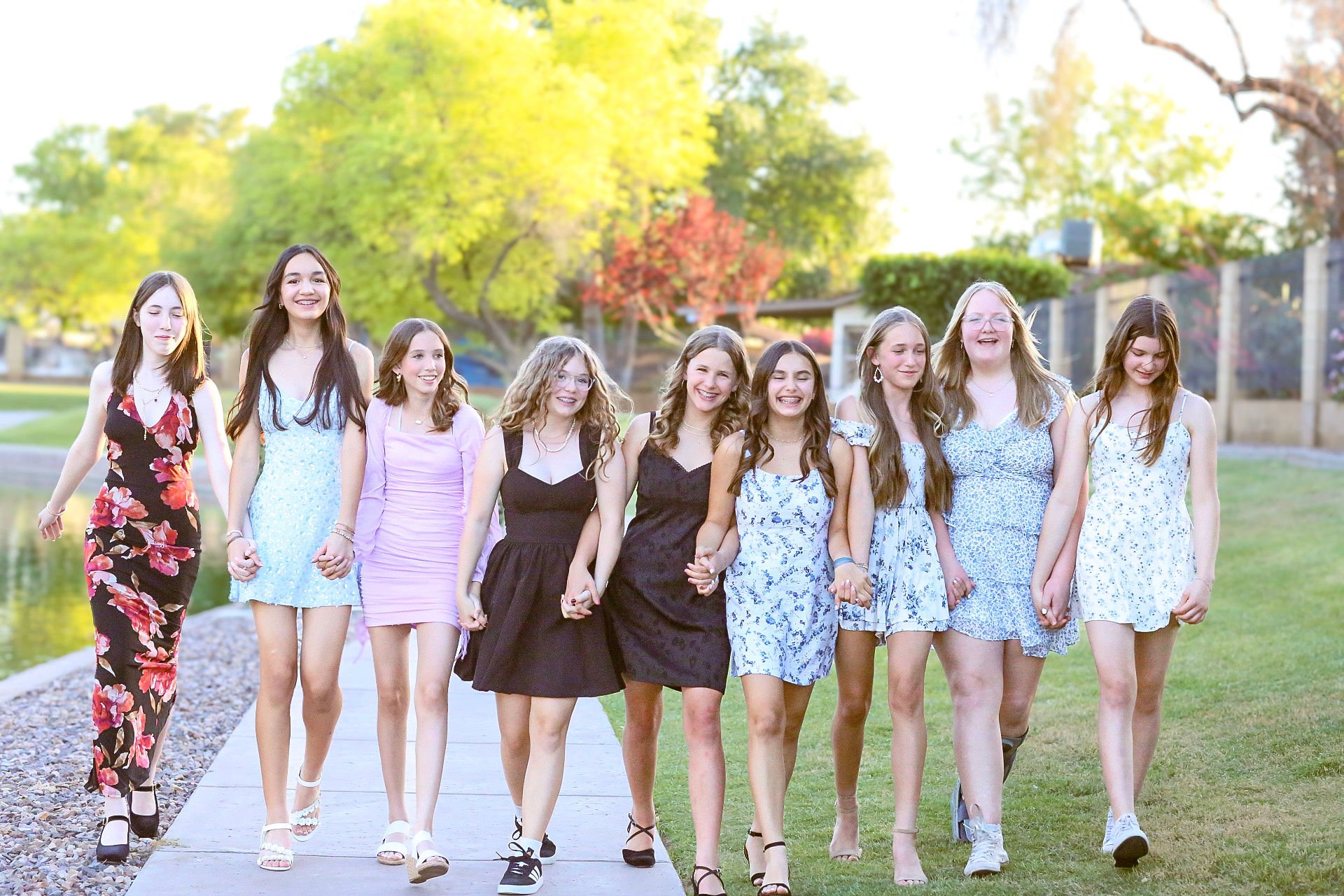 Group of ten young women walking together outdoors on a sunny day before their school dance, holding hands and smiling, with trees and a grassy area in the background.