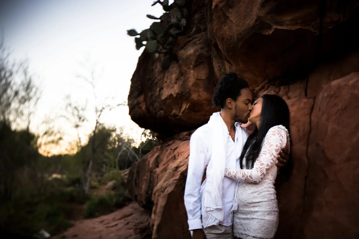 A couple sharing a kiss against a rocky cliff during sunset in a natural setting with trees in the background prior to their elopement wedding in Sedona Arizona