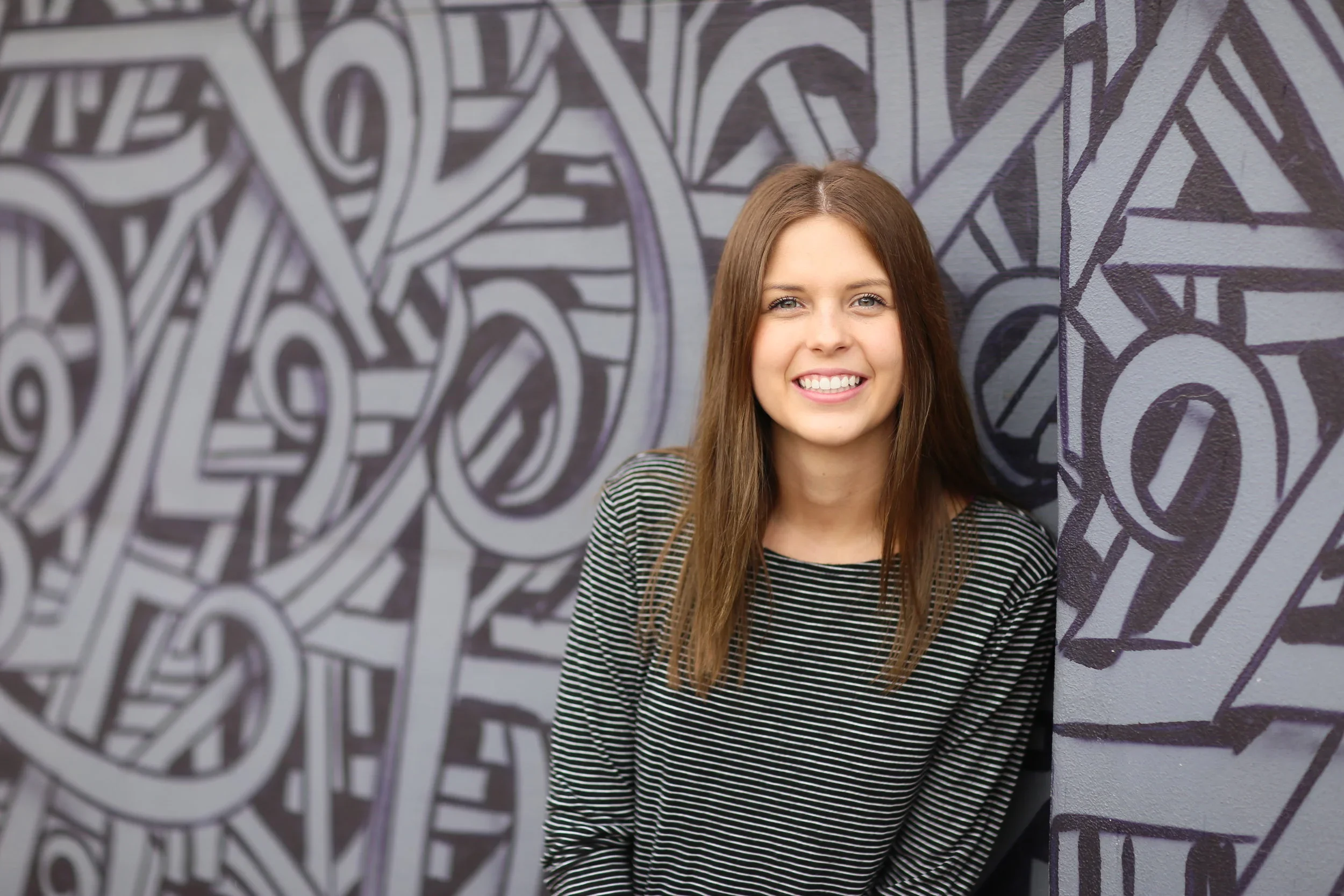A young woman with long brown hair smiling, wearing a black and white striped shirt, standing in front of a wall with abstract black and white geometric artwork for a senior photo