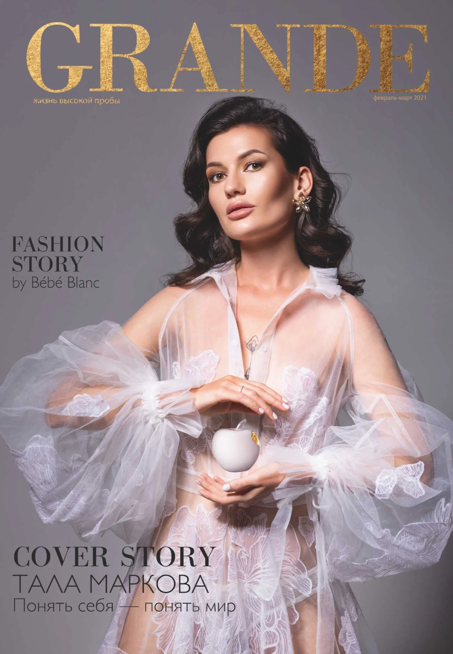 A woman with dark, wavy hair wearing a sheer, white lace dress with butterfly embroidery, holding a small white apple-shaped object.