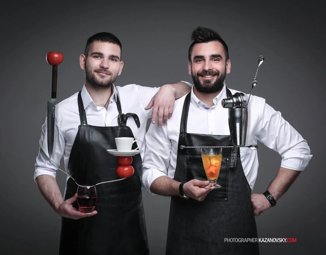Two men wearing white shirts and black aprons, posing with bar tools and drinks, against a gray background.