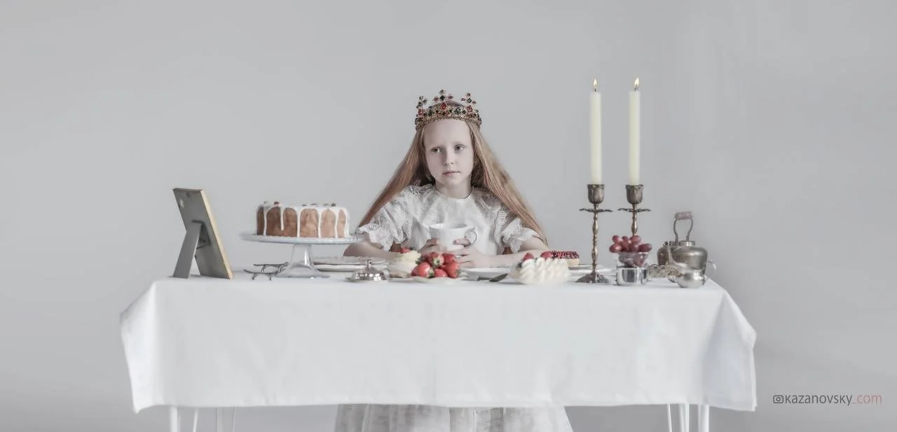 A young girl with a crown sitting at a decorated table with cake, strawberries, and candles, holding a teacup, against a plain gray background.