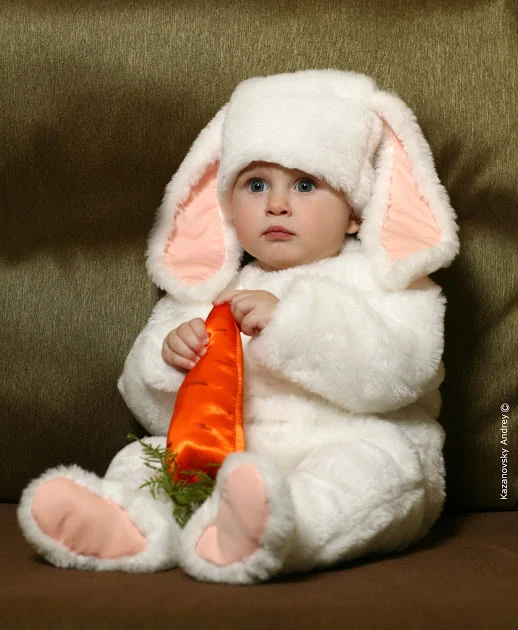 A young child dressed in a fluffy white bunny costume with pink ears, sitting on a brown sofa and holding an orange cloth
