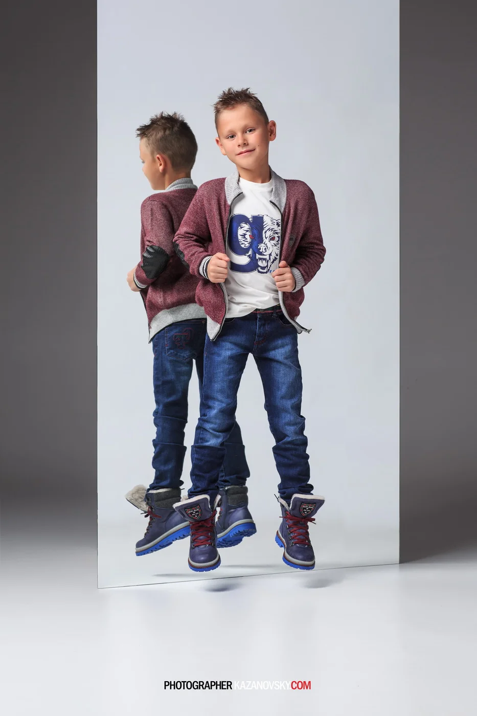 A young boy with spiky hair, wearing a white graphic t-shirt, a maroon jacket, and blue jeans, standing in front of a mirror reflection. He is wearing dark shoes with red laces and a badge on the tongue. The background is divided into a white and gra