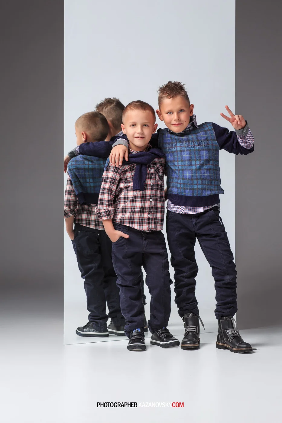 Two young boys standing in front of a large mirror, with one boy making a peace sign and the other boy with his hand in his pocket, both wearing plaid shirts, dark pants, and sneakers.