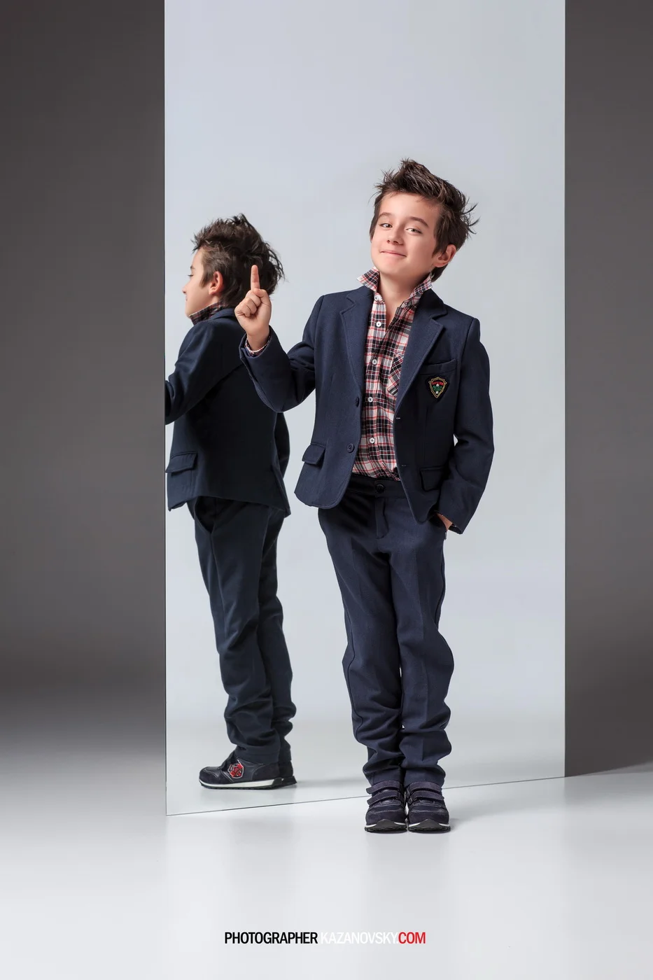 A young boy in a navy blazer and plaid shirt standing next to a mirror, with both arms in his pockets and a confident smile. The mirror reflects his back, showing the same outfit. The background is a plain, light-colored studio setting.