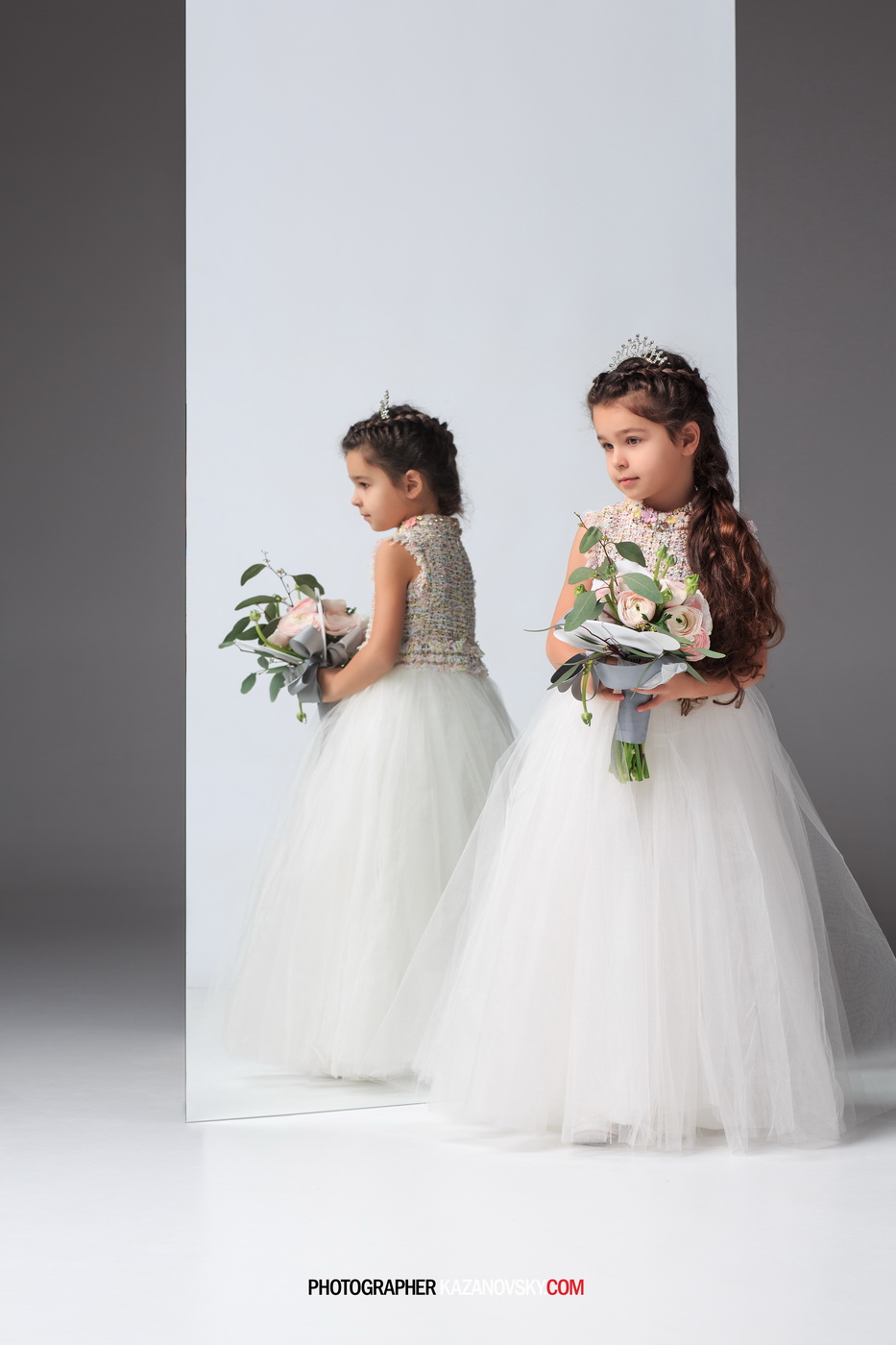 Two young girls in white dresses and tiaras holding bouquets of pink flowers, standing in front of a mirror. One girl faces the mirror while the other portrays her reflection.