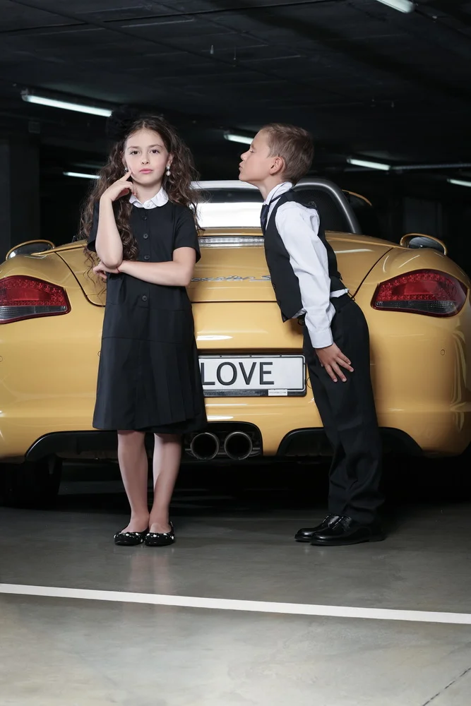 A girl and boy in formal attire standing in front of a yellow sports car in a parking garage, with the car's license plate displaying the word 'LOVE'.