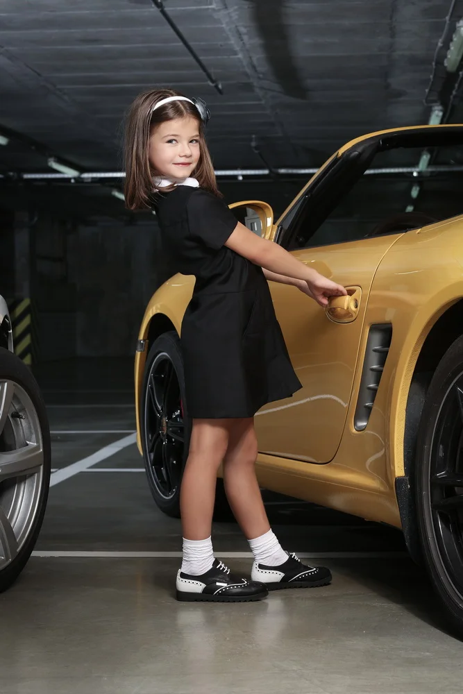 Young girl with brown hair, wearing a black dress, white socks, and black and white shoes, standing next to a yellow sports car in a parking garage, holding the door handle.
