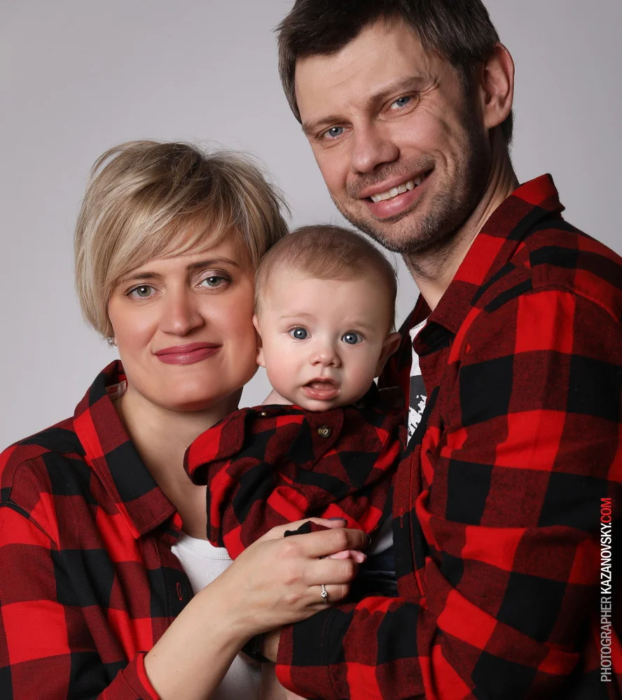 Family portrait of a woman, man, and baby all wearing red and black plaid shirts, smiling and looking at the camera against a plain background.