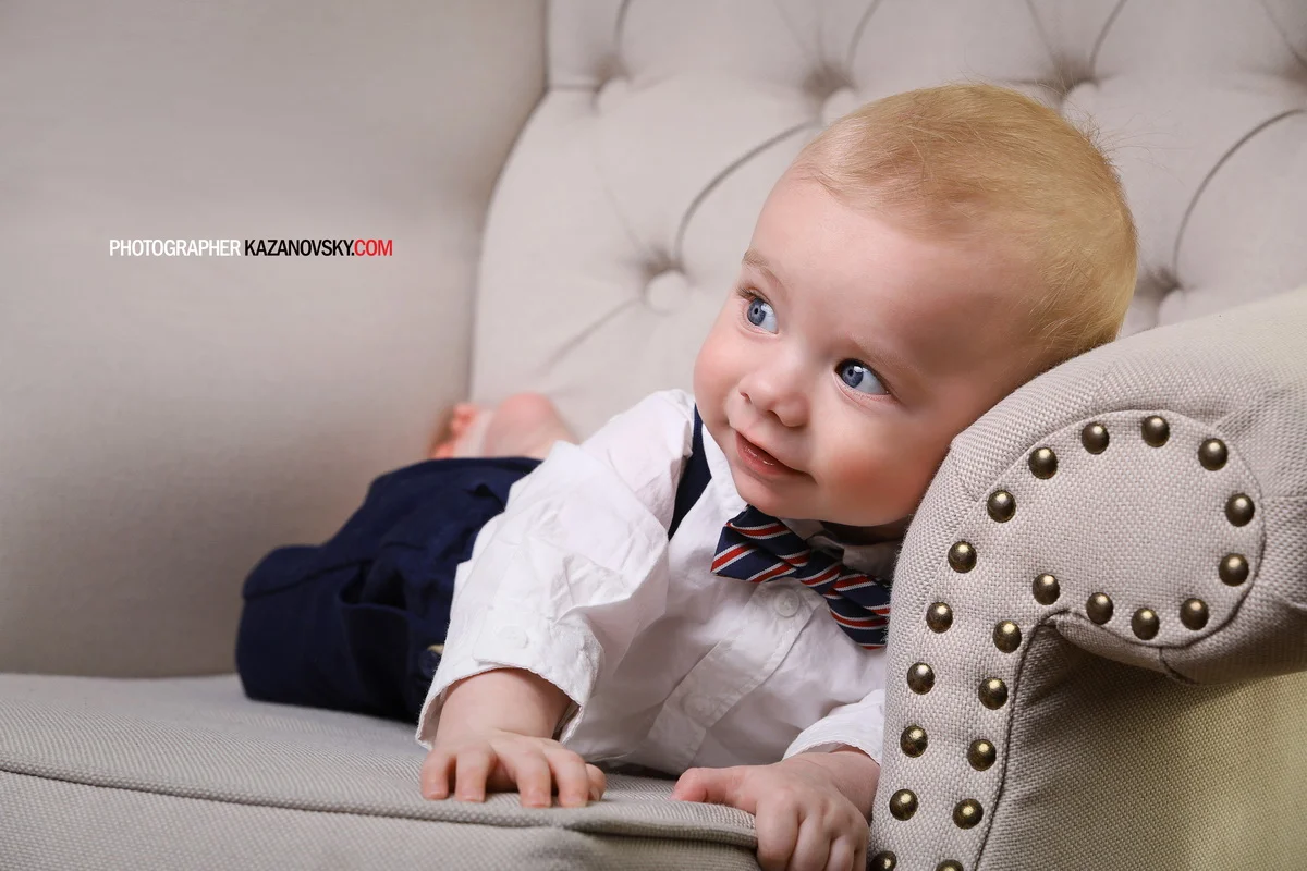Young boy with blonde hair and blue eyes lying on a beige tufted sofa, dressed in a white shirt, navy pants, and a striped bow tie, looking to the side with a slight smile.