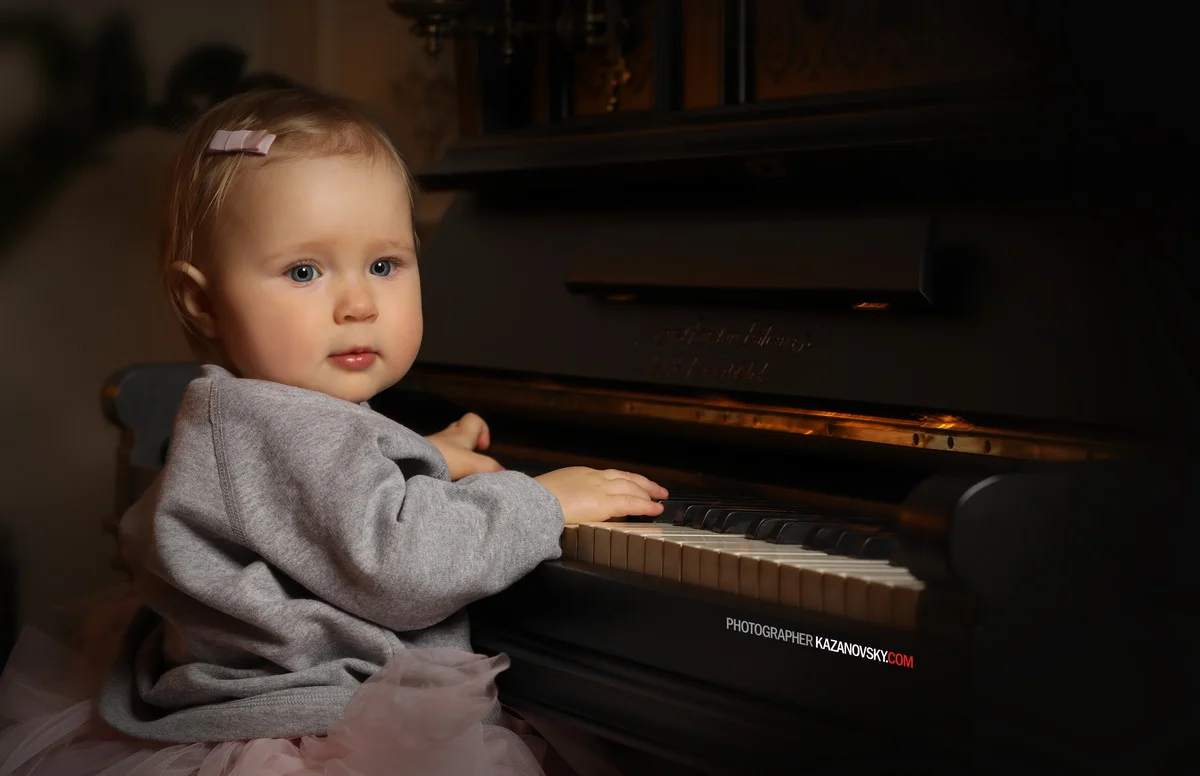 Young girl with blonde hair and a pink hair clip, wearing a gray sweatshirt, sitting at a black piano, looking to her left.