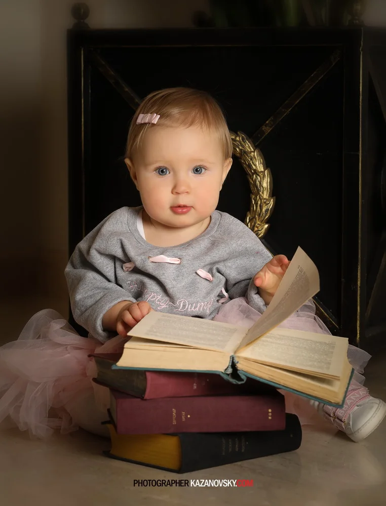 A young girl with blue eyes, light brown hair with a pink hair clip, sitting on the floor with several books, looking at the camera. She is wearing a grey top with pink bows and a pink tulle skirt. There is a black fireplace with a gold decorative wr