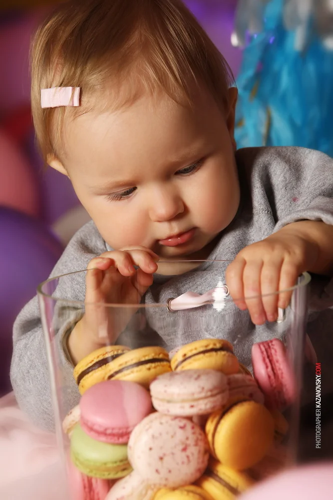 Young child with a pink hair clip looking at a glass container filled with colorful macarons and pink candies.