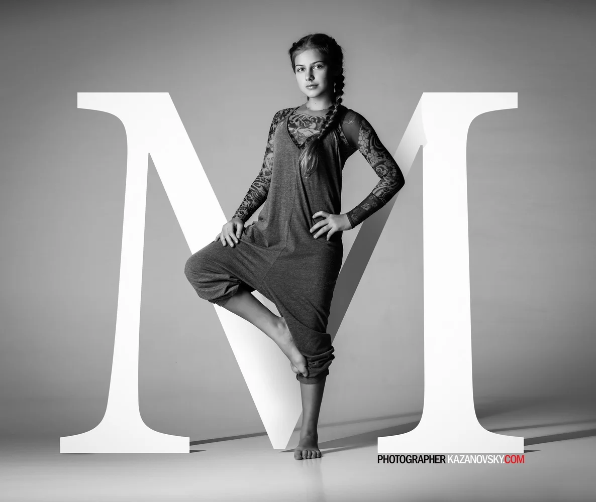 A young woman standing inside a large letter 'V' in front of a plain background, posing confidently with one foot on the ground and the other knee resting on the letter, wearing a long-sleeved lace top and loose pants, in a black-and-white studio pho