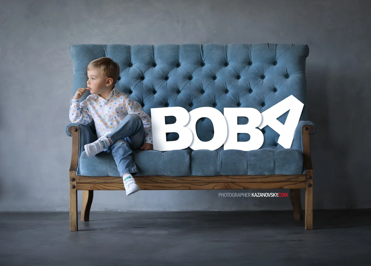 A young boy sits on a blue vintage-style sofa with a tufted back. There are large 3D white letters spelling 'BOBA' placed on the sofa beside him. The boy is wearing a colorful polka dot shirt, jeans, and white socks, and is looking to the side with h