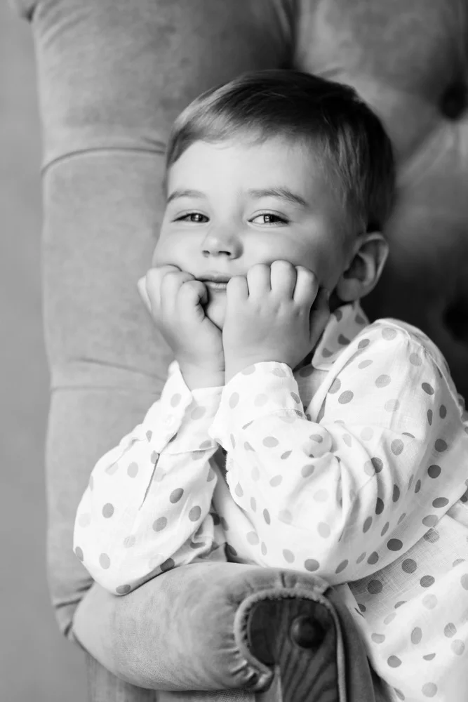 Smiling young boy with hands on cheeks, sitting on a plush armchair, wearing a polka dot shirt.