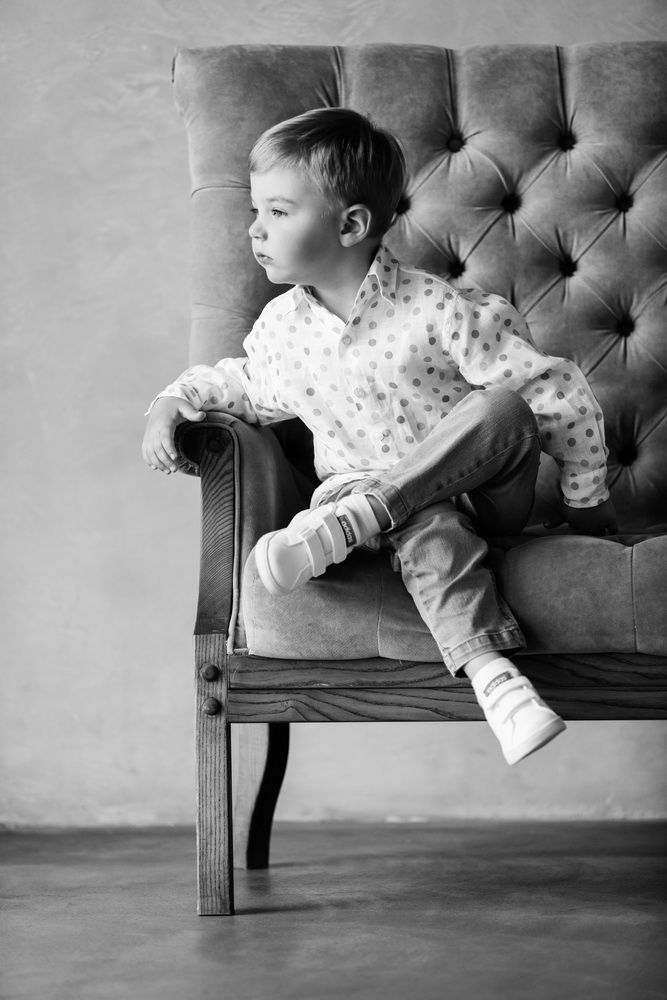 A young boy with light hair, wearing a polka dot shirt, jeans, and sneakers, sitting sideways on a vintage upholstered armchair and gazing thoughtfully to the left.