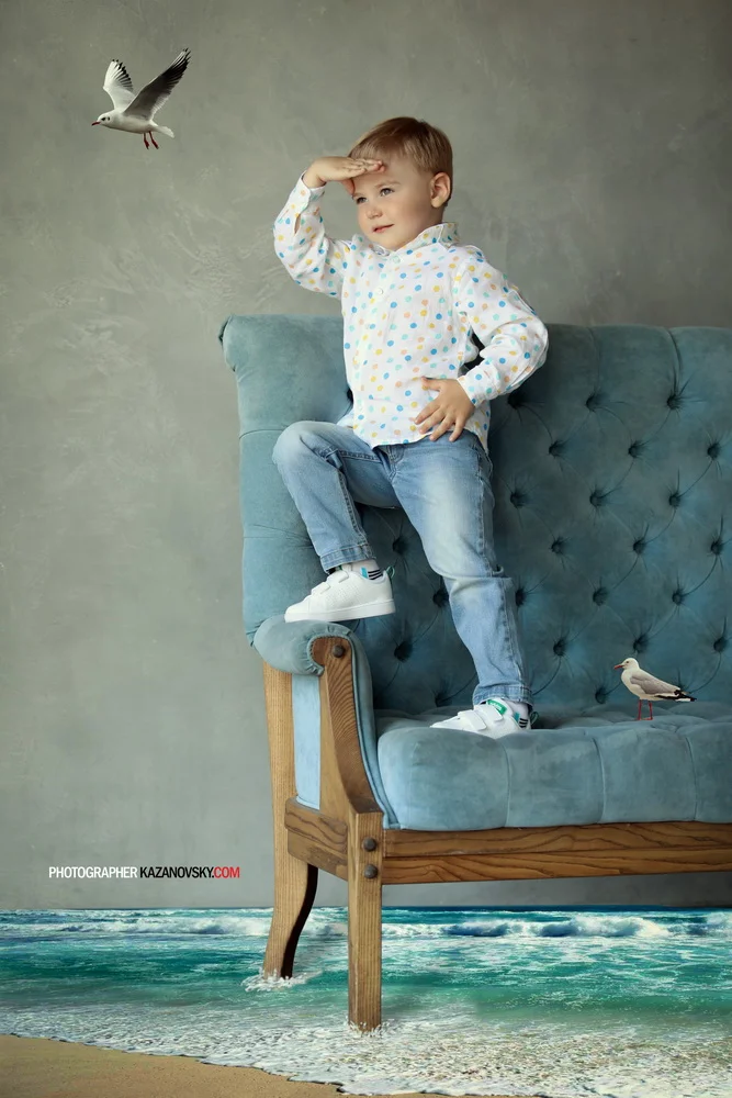 A boy sitting on a teal tufted couch with a background that appears as a beach scene, with two seagulls—one flying overhead and one standing on the couch—creating an illusion of a beach setting.