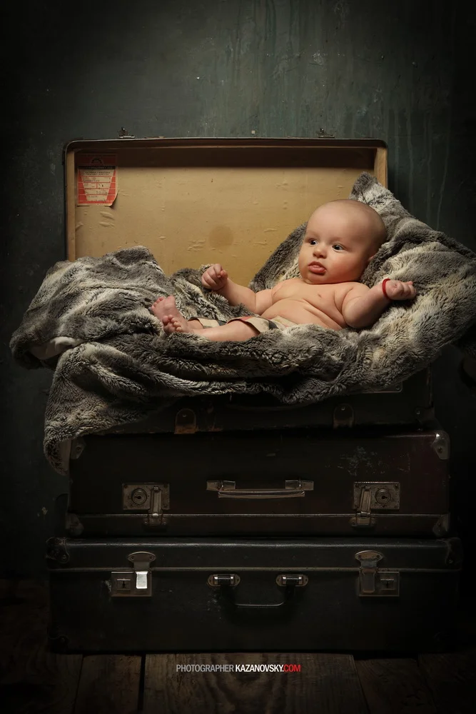 A baby lying on a soft, plush blanket inside an open vintage suitcase against a dark wall.