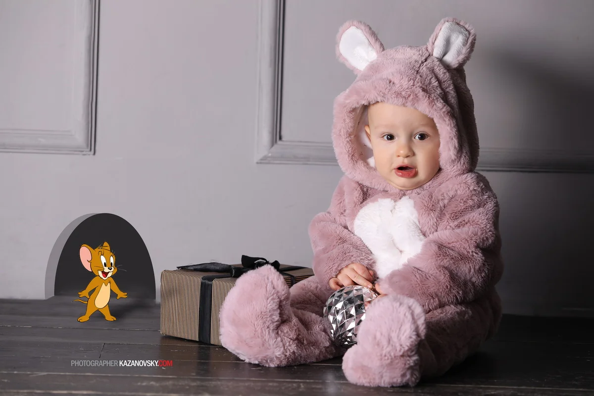 A baby in a pink, fluffy animal costume with ears, sitting on the floor holding a shiny silver ornament. There is a striped gift box nearby and cartoon character Jerry the mouse from Tom and Jerry near a small faux fireplace.