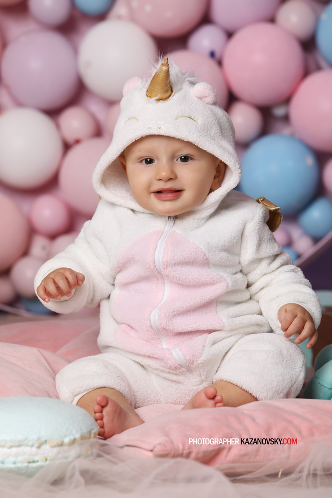 Child dressed in a unicorn onesie with a hood featuring a golden horn, sitting on pink and blue cushions with a background of pastel-colored balloons.