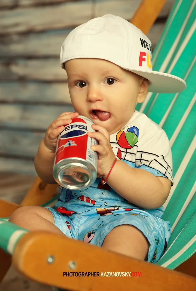 A young child sitting in a green and white striped chair, wearing a white cap tilted sideways with the text "WE F" visible, holding and about to drink a can of Pepsi. The child is dressed in a colorful summer outfit with cartoon patterns.