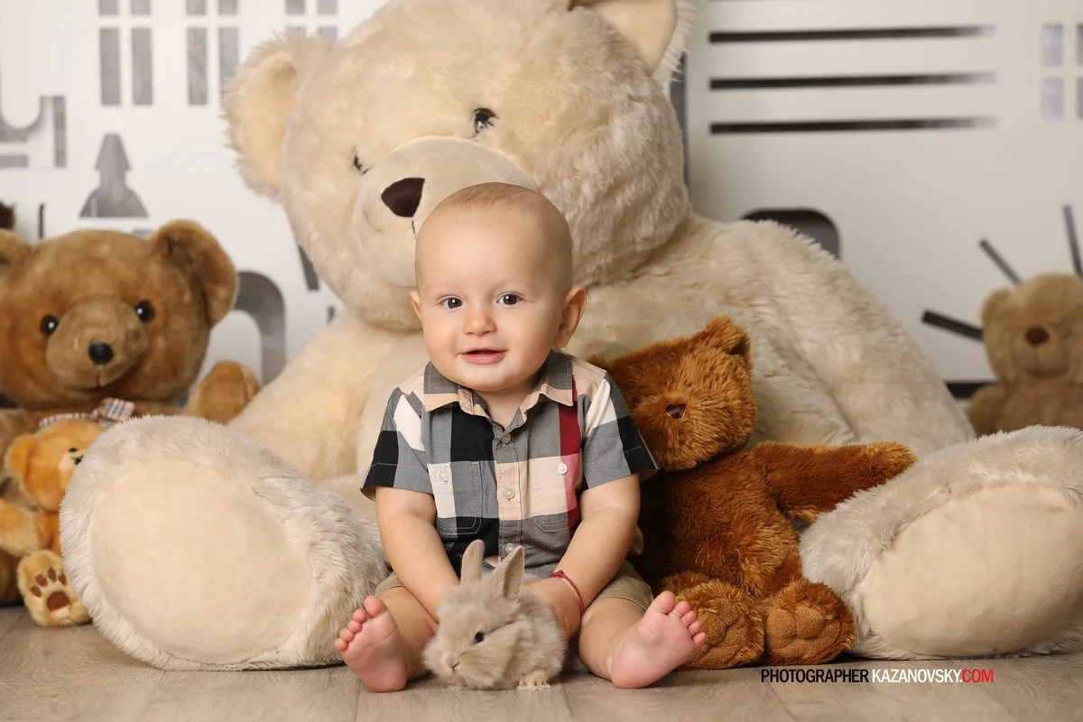 A young boy sitting on the floor surrounded by teddy bears and holding a stuffed rabbit, with a large teddy bear behind him.