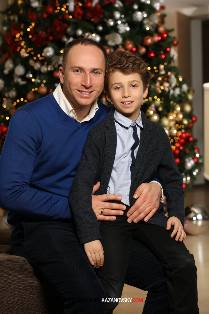 A man and a young boy sitting together, smiling, with a decorated Christmas tree in the background.