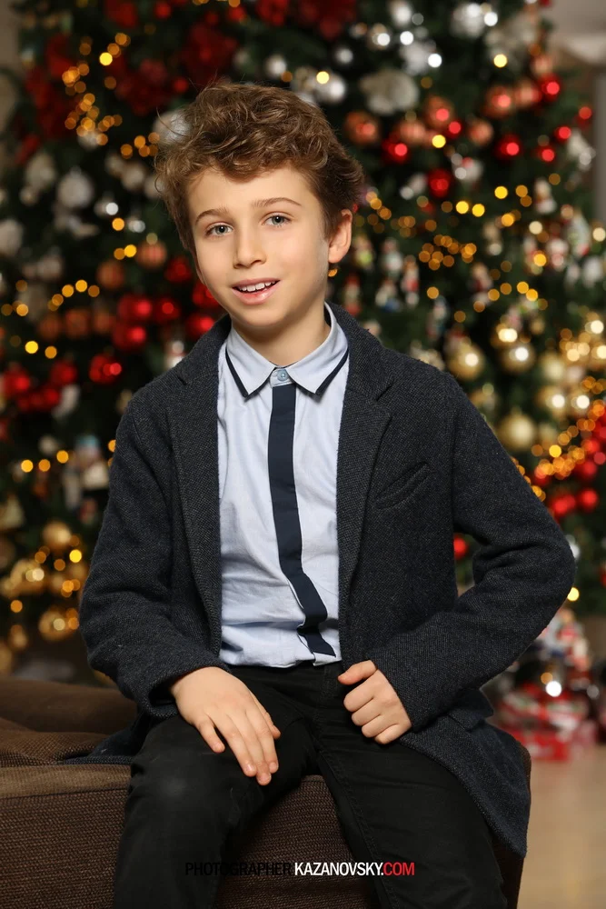 A young boy with light brown, curly hair and a fair complexion sitting on a brown bench in front of a decorated Christmas tree with red, white, gold, and silver ornaments, and warm white lights, smiling at the camera. The boy is wearing a light blue 