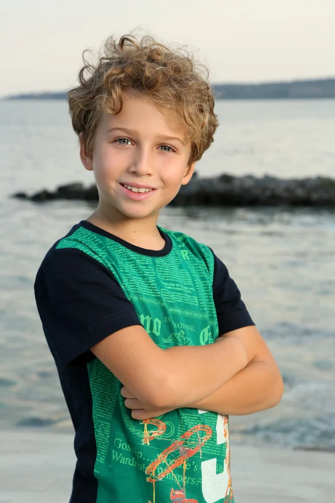 Young boy with curly blonde hair and blue eyes standing on the beach with arms crossed, smiling at the camera, with water and rocks in the background.