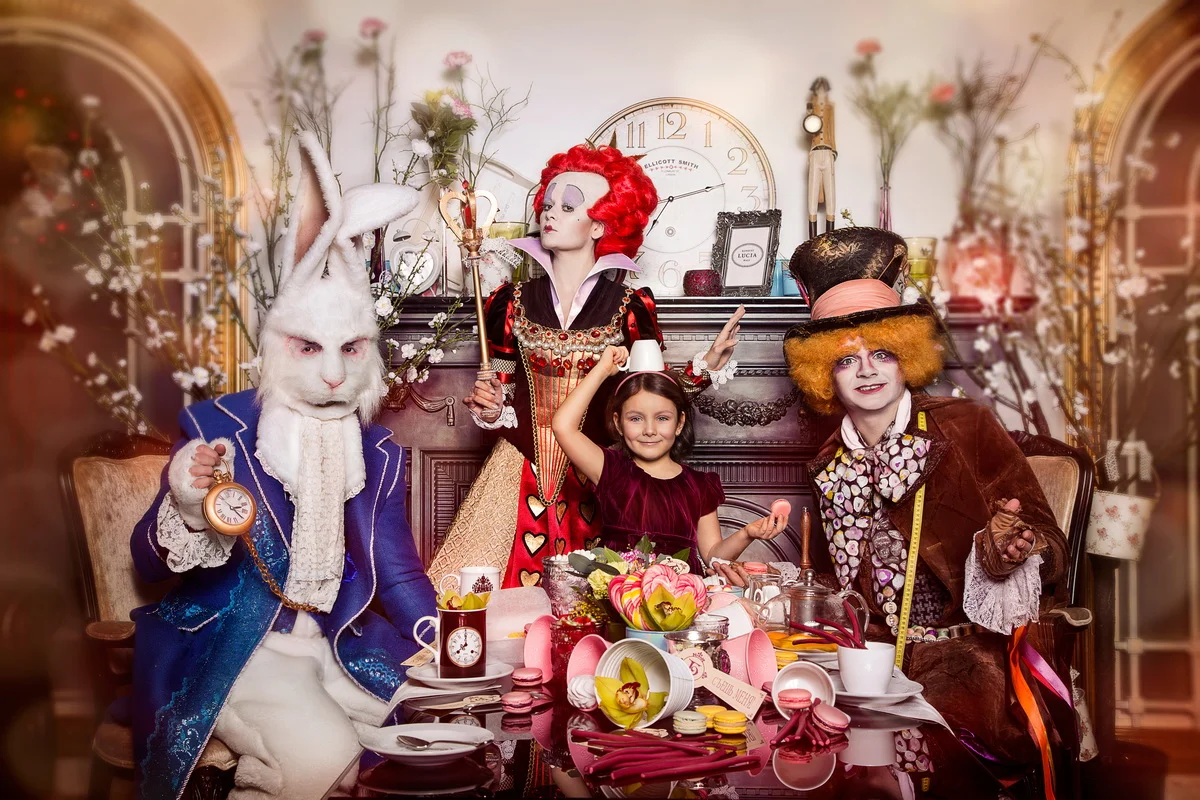 People in costumes gathered around a decorated table with food and drinks, with a woman dressed as the Queen of Hearts, a person in a rabbit costume, and a person as the Mad Hatter from Alice in Wonderland, in a vintage-style room.
