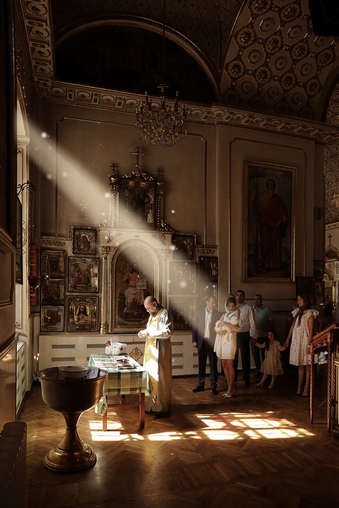 A priest officiates a baptism inside a church, with visitors observing. Sunlight streams through windows, illuminating religious icons and ornate interior decor.