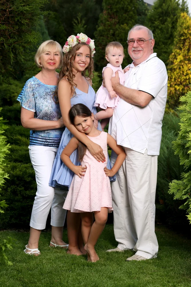 A multigeneration family of five stands outdoors on green grass surrounded by lush trees. An older woman with short blonde hair wears a blue tie-dye blouse and white pants. A young woman with long curly hair and a flower crown wears a light blue dres