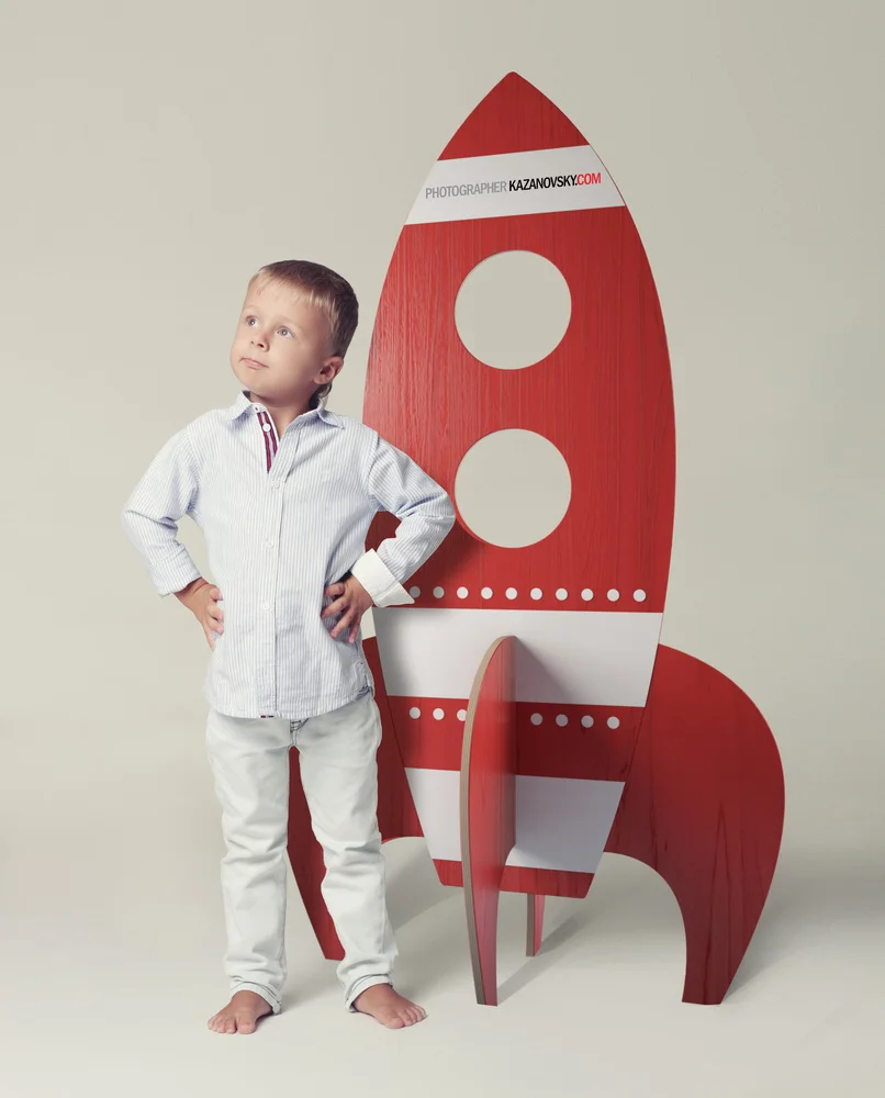 A young boy in a striped button-up shirt and white pants standing with hands on hips next to a red and white rocket-shaped shelf.