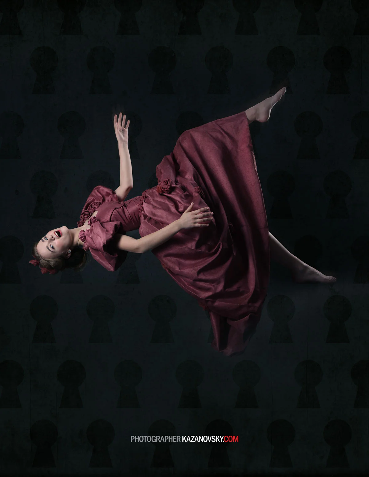 A young girl in a maroon Victorian-style dress lying on her side against a dark background with keyhole patterns, smiling and looking up, with her right arm raised and her left arm resting on her dress.