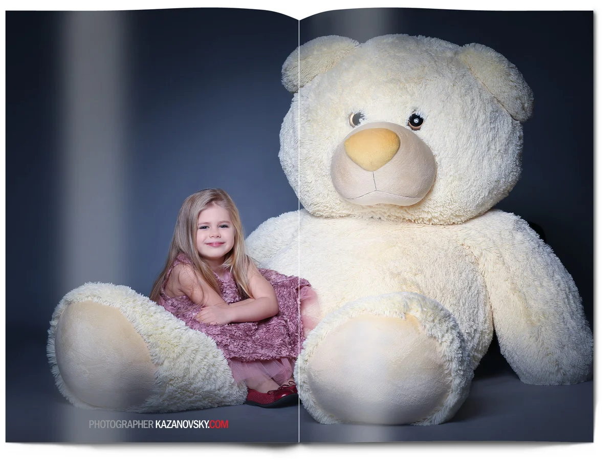 A young girl in a pink dress sitting next to a large white teddy bear against a dark background.