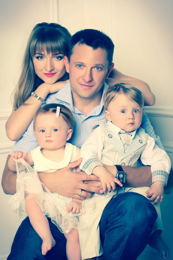A family portrait of a man, woman, and two young children indoors with a light-colored background.
