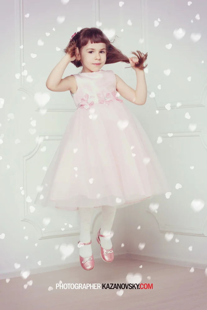 A young girl in a pink dress and pink shoes is jumping with a background of white wall paneling and white floating heart-shaped lights.