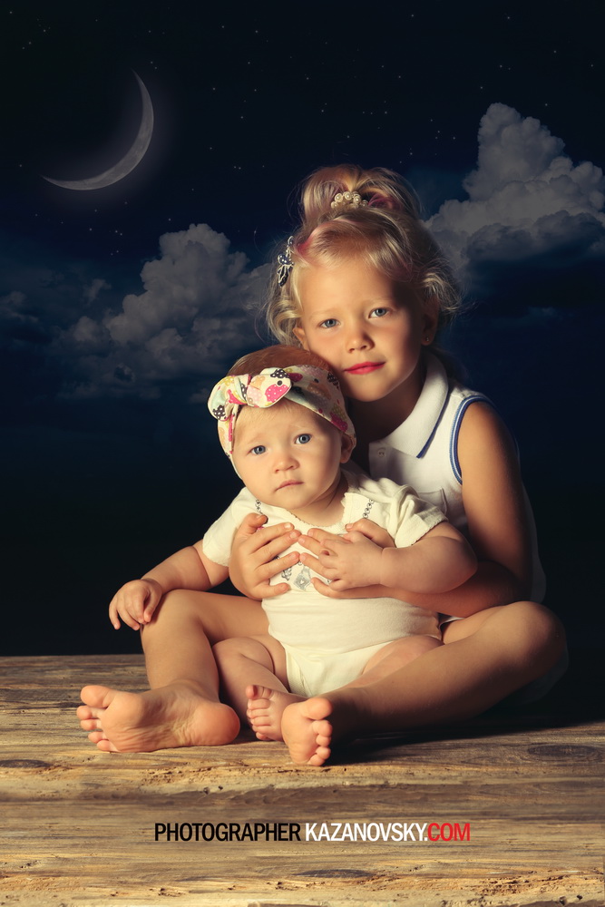 A young girl and a baby sitting on a wooden surface outdoors at night, with a starry sky, crescent moon, and clouds in the background.