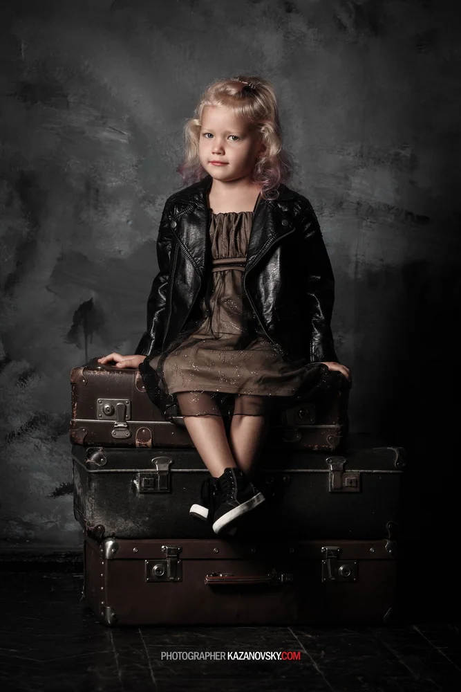 A young girl with blonde hair, dressed in a brown dress and black sneakers, sitting on vintage suitcases against a dark textured background.