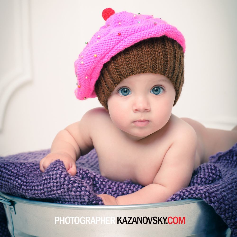 A baby with bright blue eyes and fair skin lying on a purple textured blanket, wearing a pink knitted hat decorated with multicolored beads and a brown beanie underneath, in a metal basin. The background is plain and light-colored.