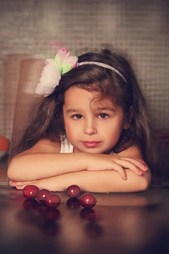 Young girl with long dark hair and a large pink and white fabric flower headband resting her chin on her folded arms on a dark wooden table with red grapes in front of her.