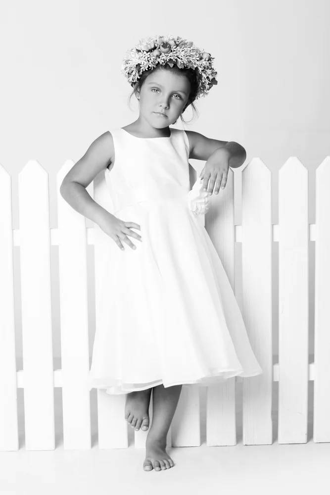A young girl in a white dress and a flower crown standing barefoot in front of a white picket fence, posing with one hand on her hip and the other resting on the fence.
