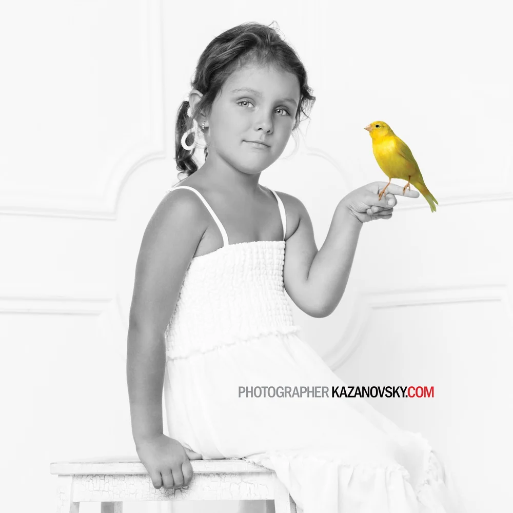 Black and white photo of a young girl sitting on a stool, holding a yellow bird on her finger, with the girl and bird in color.